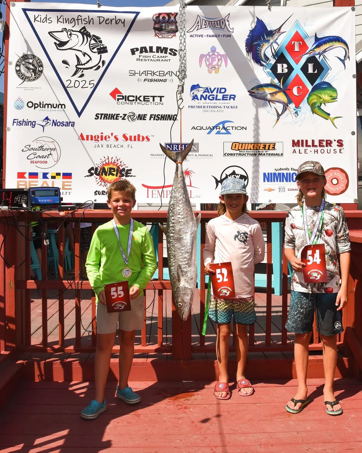 Three young anglers with tournament numbers 55, 52, and 54 posing at the Kids Kingfish Derby 2025 awards banner