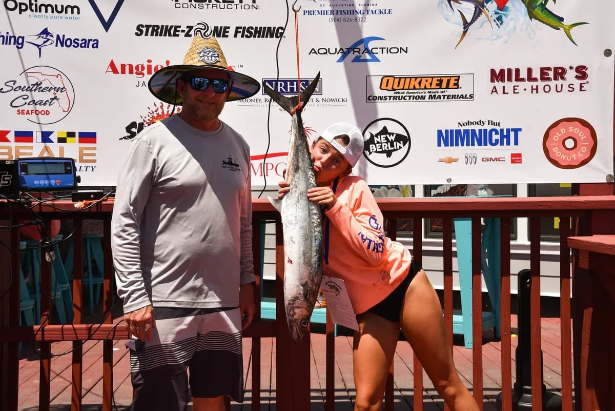 Young female angler holding a large Kingfish at the official weigh-in banner beside a tournament official