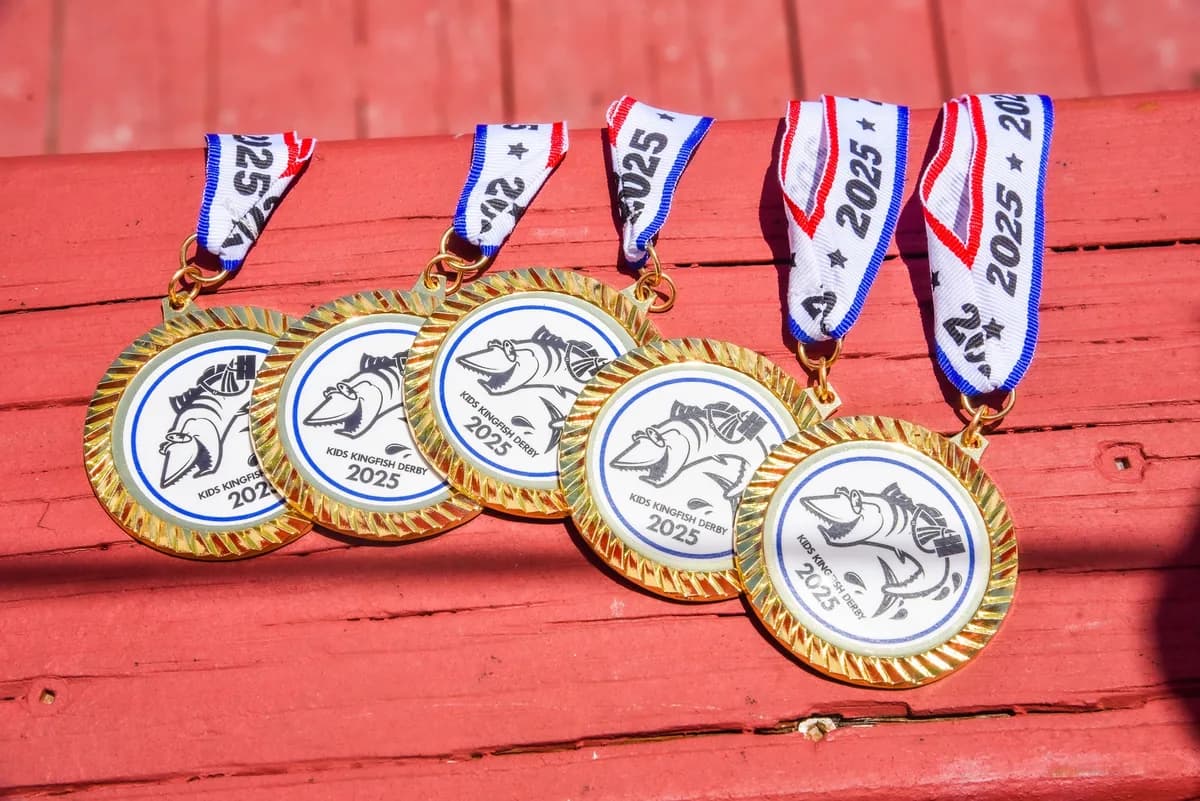 Five 2025 Kids Kingfish Derby gold medals with red, white, and blue ribbons displayed on a red wooden railing