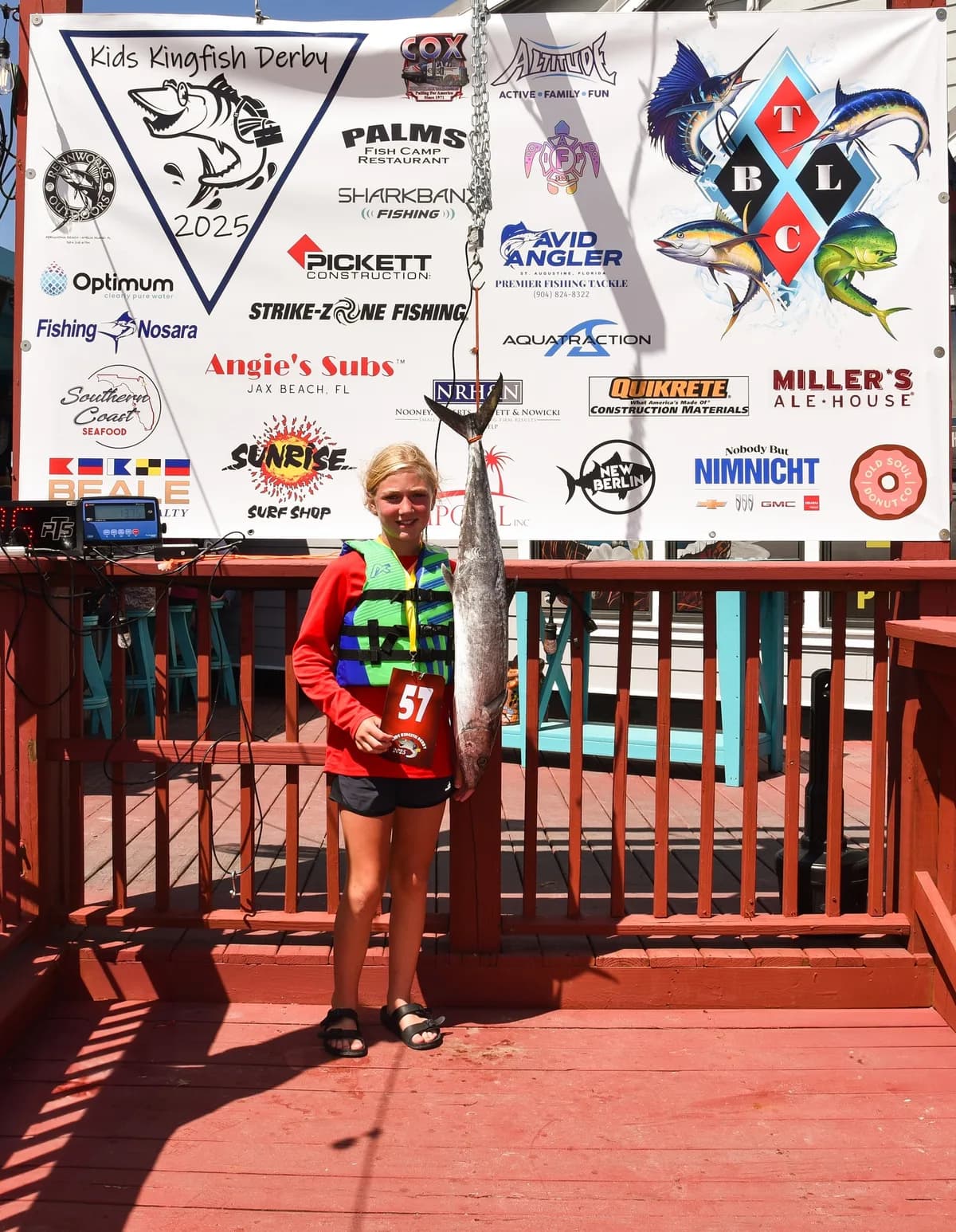 Young angler with tournament number 57 holding a full-length Kingfish at the Kids Kingfish Derby 2025 weigh-in banner